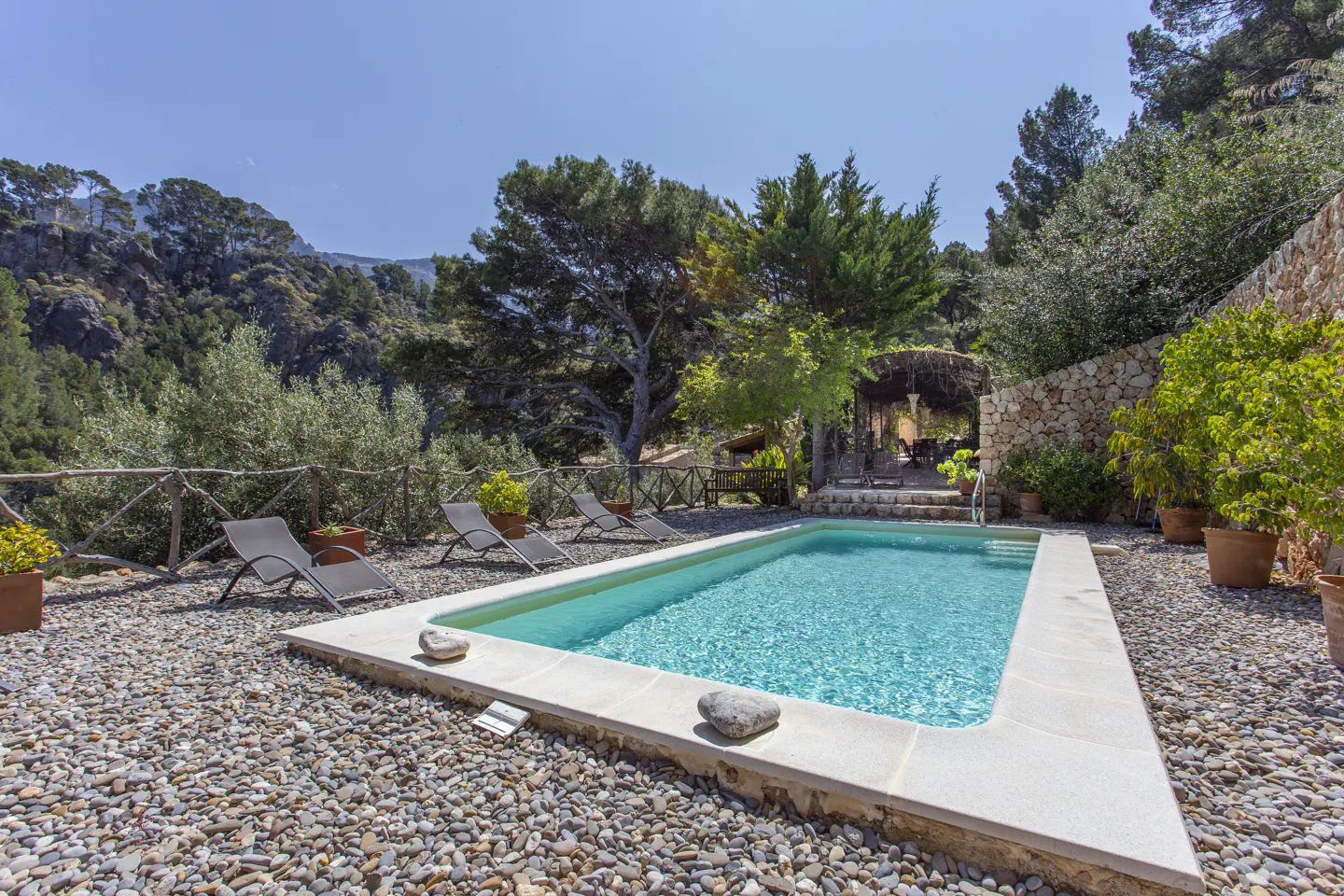 Outdoor pool with turquoise water, surrounded by stone, lounge chairs, and lush greenery. A stone wall and trees form the backdrop.
