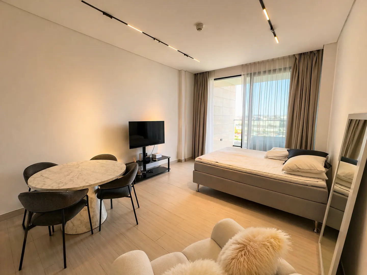 A modern studio apartment with a bed, dining table, TV, and a large window with curtains. The room is bright and airy with light wood floors.