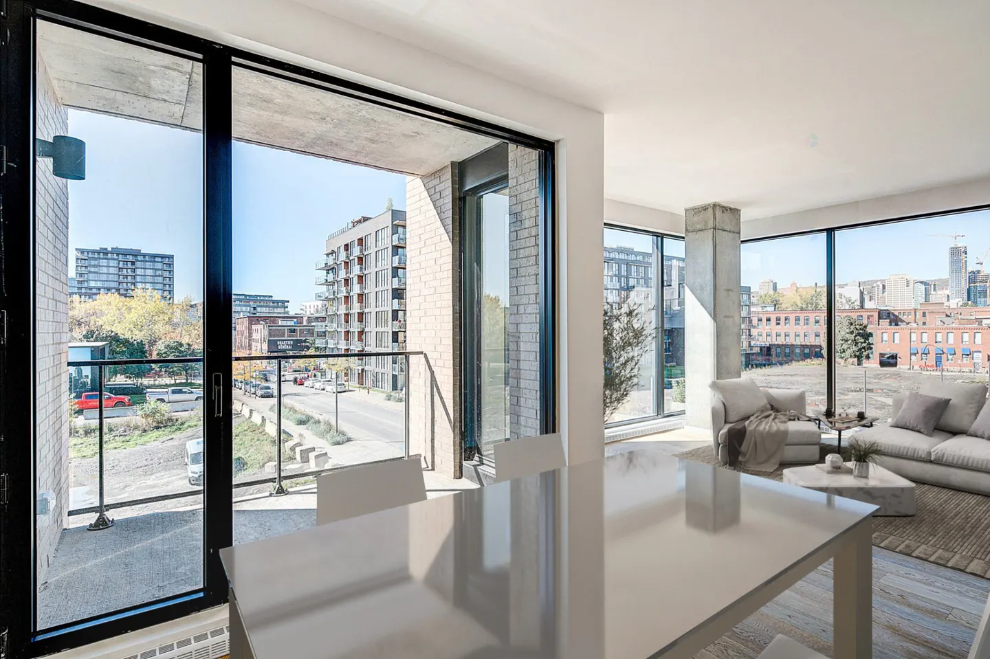 Bright modern living room with a white table, sofa, and large windows showing a city view. Balcony with black railings is visible.