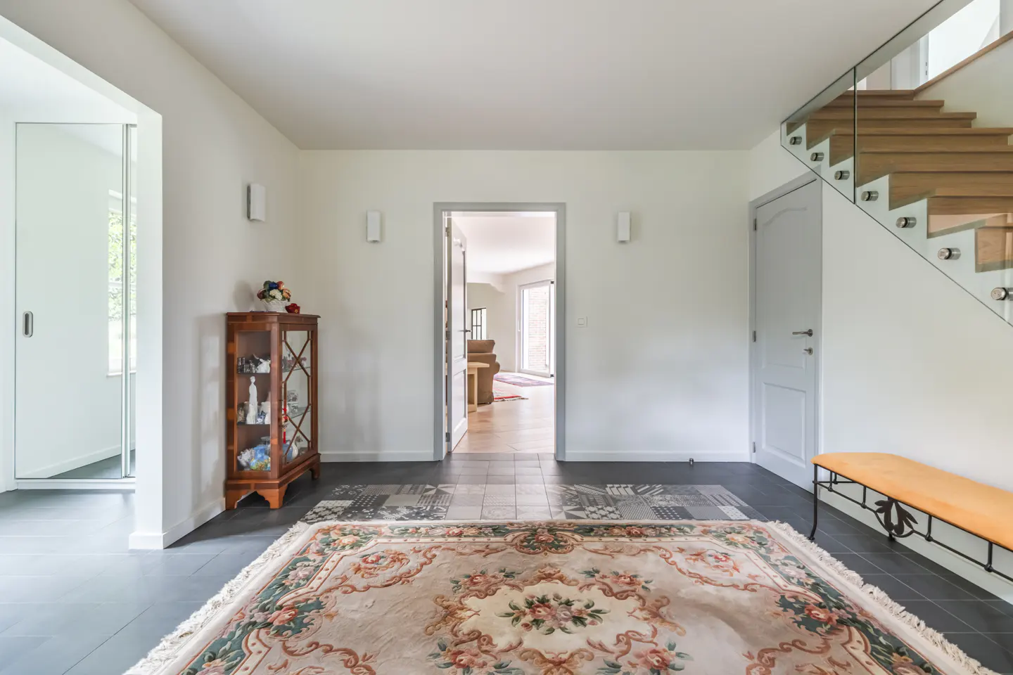 Bright foyer with a floral rug, a wooden cabinet, and a staircase with glass railings. A doorway leads to another room.