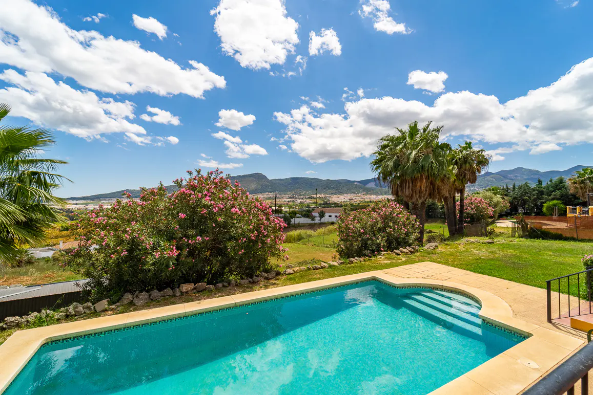 A turquoise pool with steps, surrounded by green grass, pink flowers, and palm trees under a blue, cloudy sky.