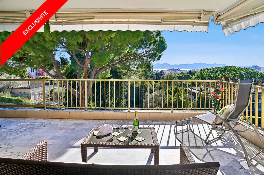 Balcony view with gold railing, table set with champagne, glasses, and hat. Lounge chair and wicker furniture. Green trees and blue sky.