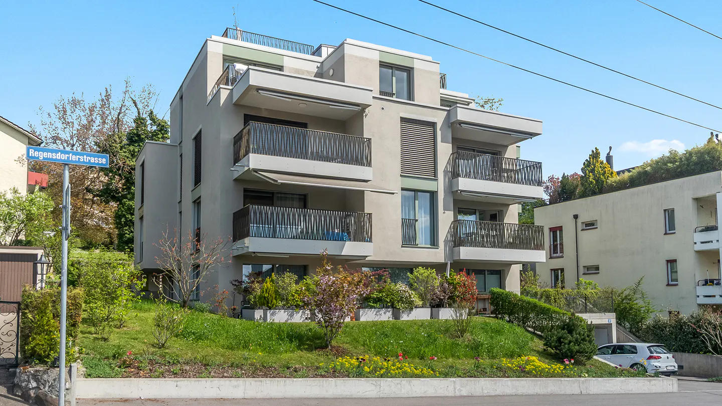 Modern apartment building with balconies, set on a grassy hill with flowers. Street sign visible.