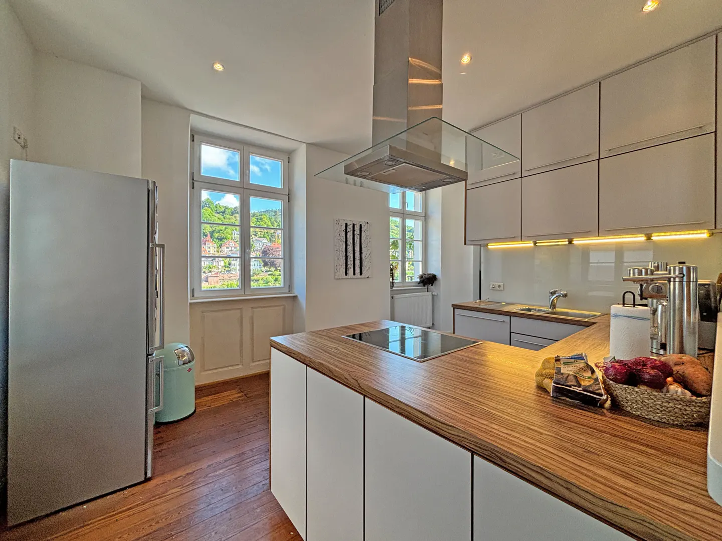 Bright kitchen with wood floors, white cabinets, and a wood countertop island. A stainless steel fridge and hood are visible. A window shows a view of a town.