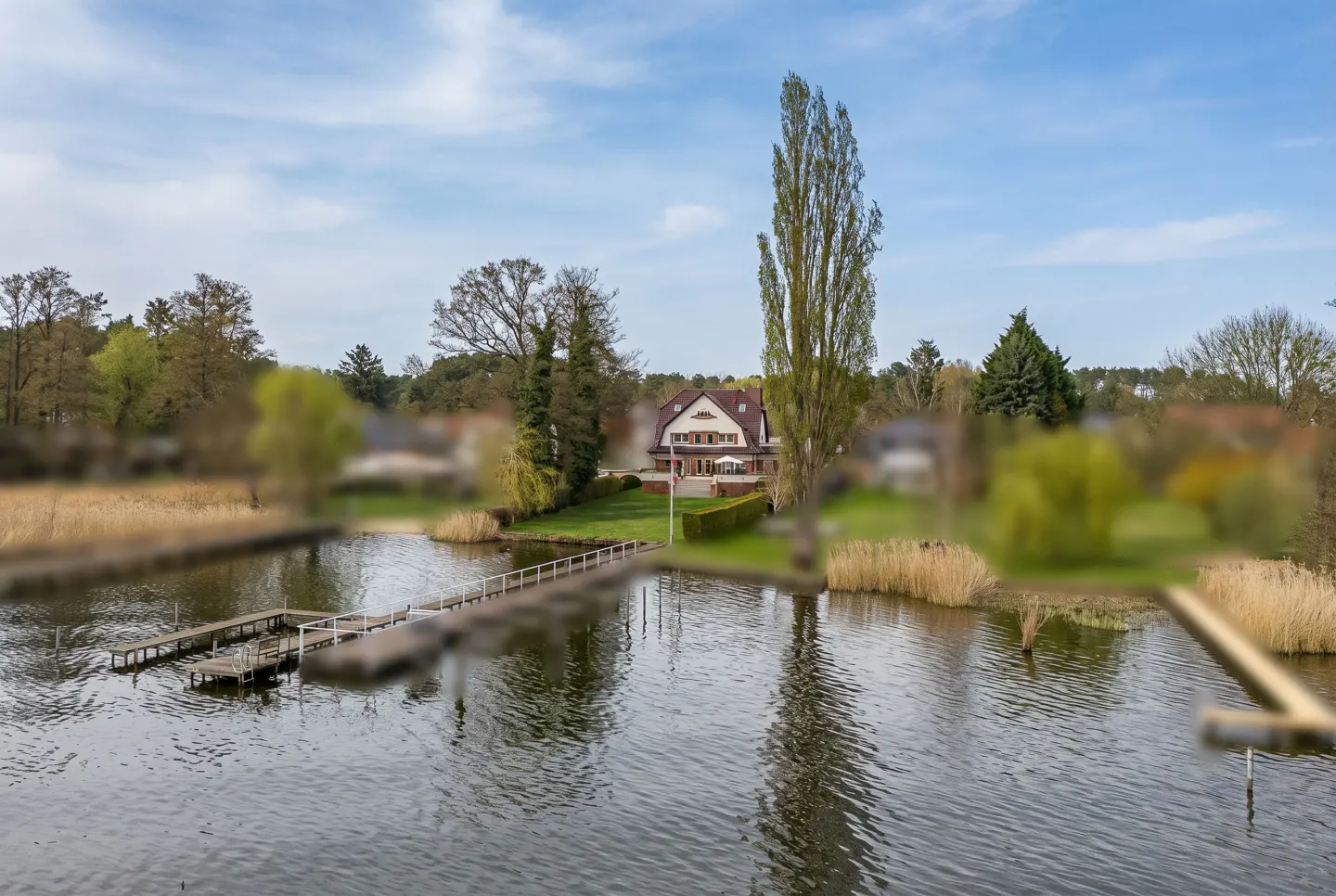 Aerial view of a lake house with a wooden dock extending into the water, surrounded by trees and reeds under a blue sky.