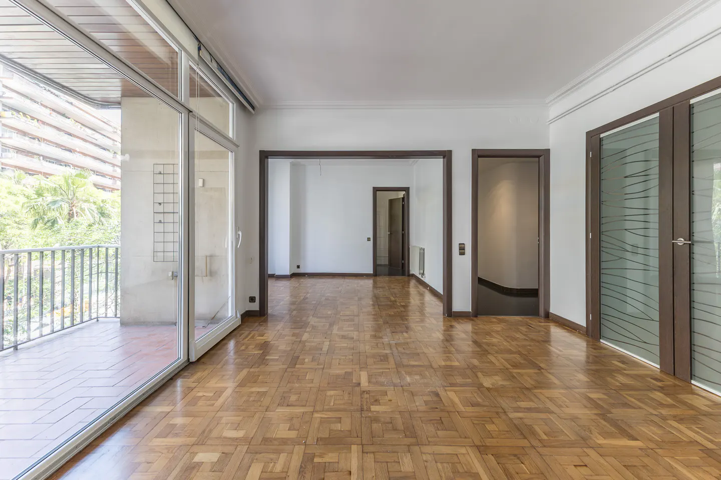 An empty room with parquet floors, white walls, and a balcony with sliding glass doors.