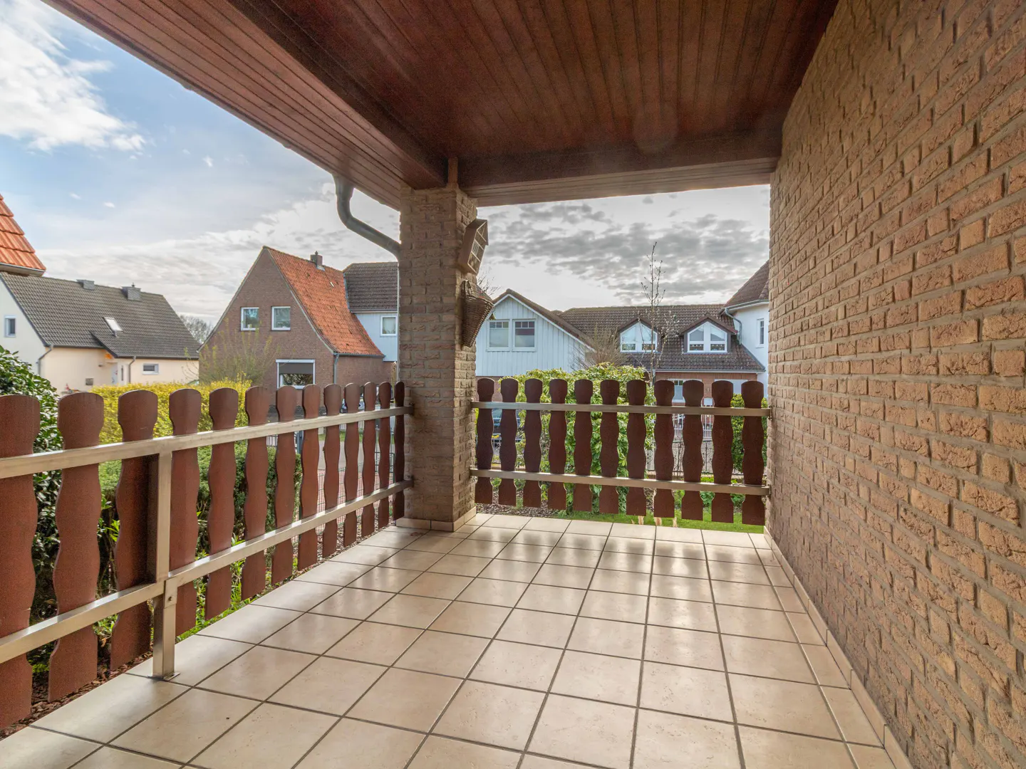 A covered porch with a brown wood ceiling, brick walls, and a brown picket fence overlooking houses.