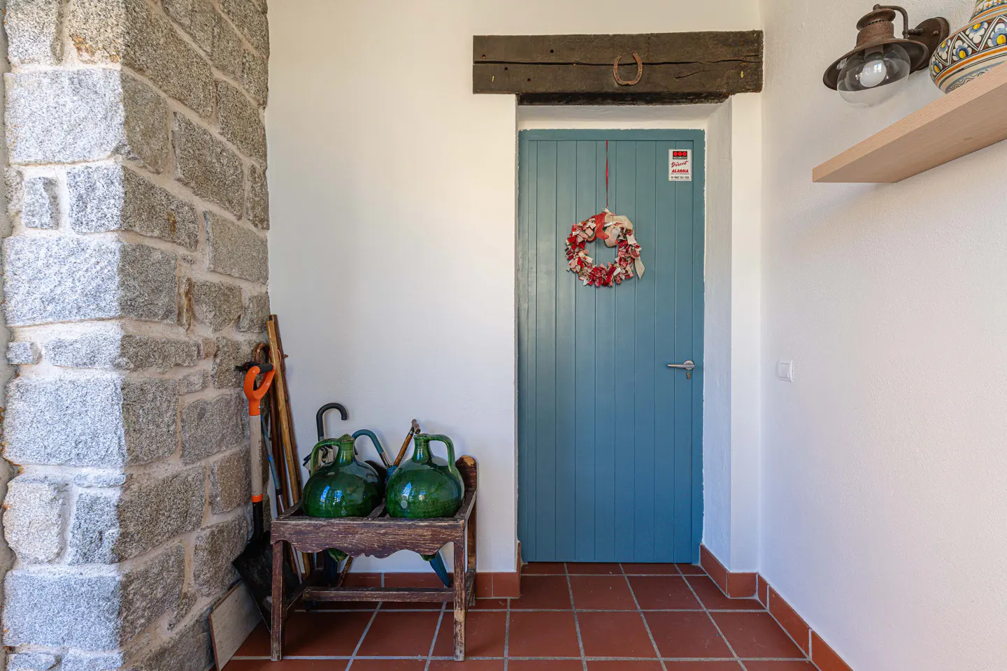 Entryway with stone wall, terracotta tile floor, and blue door with wreath. Green jugs sit on a wooden bench.