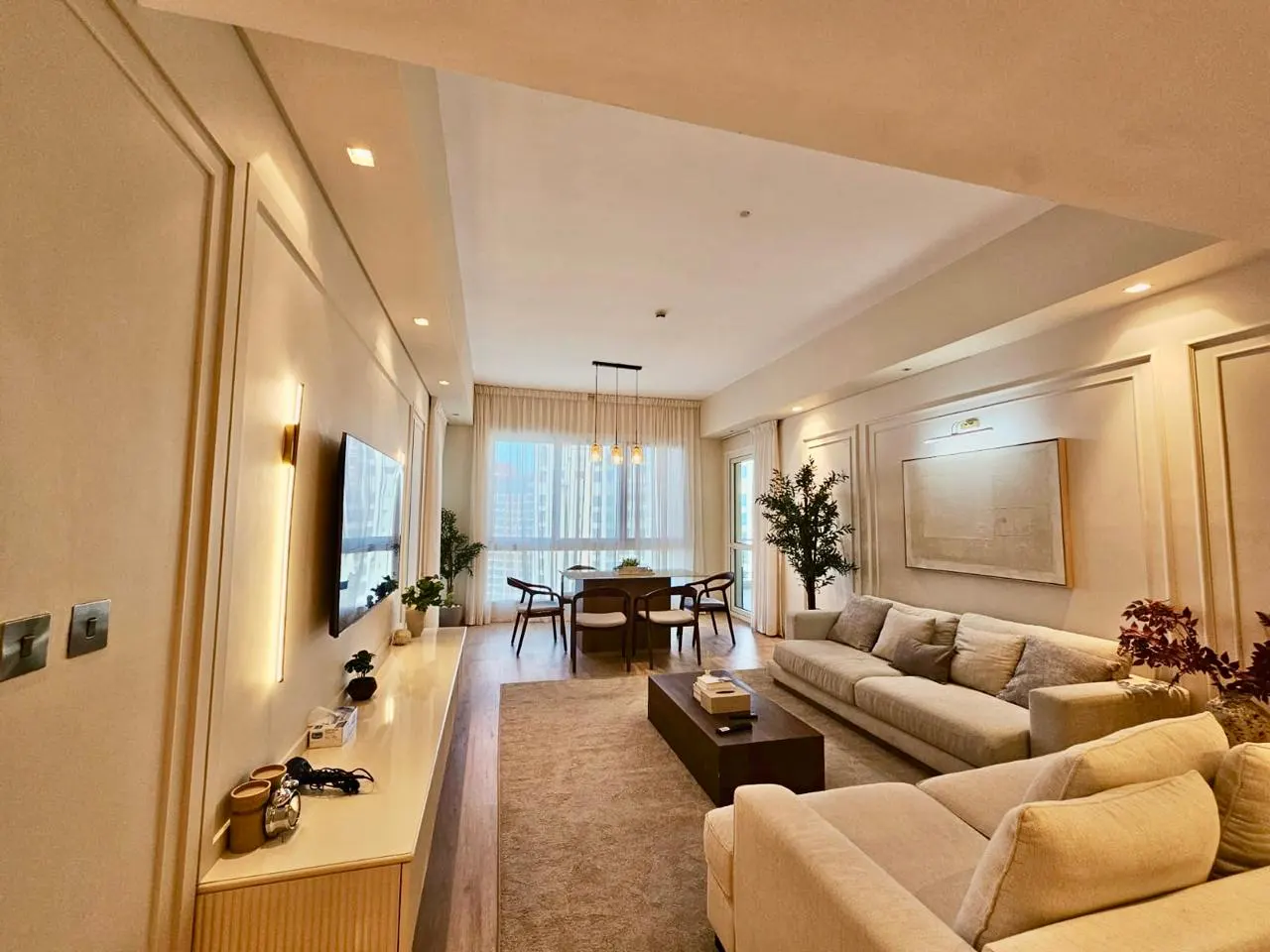 A bright, modern living room with a beige sectional sofa, a dark wood coffee table, and a dining table with chairs. Large windows offer a city view.
