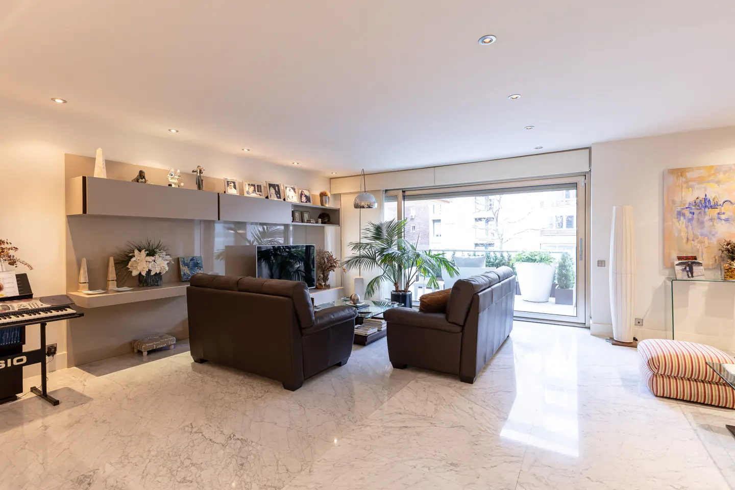 Modern living room with marble floors, two brown leather sofas, a keyboard, and a balcony view through large glass doors.