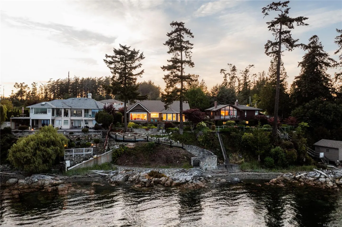 Waterfront homes on a rocky shore, with tall trees and a sunset sky.