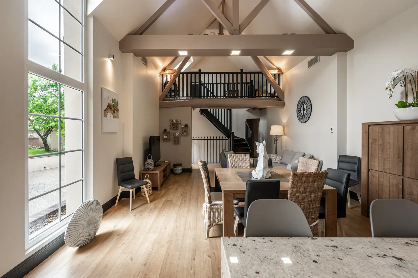 Bright, open-plan living space with wooden beams, a dining table, and a mezzanine level with black railings. Large window on the left.