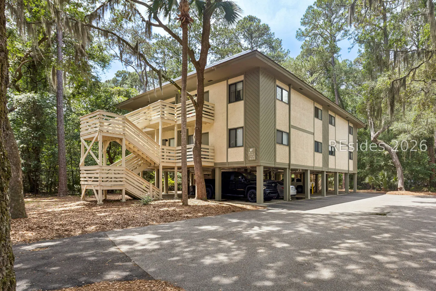 Two-story beige building on stilts with wooden stairs and balconies, surrounded by trees. Cars are parked underneath the building.