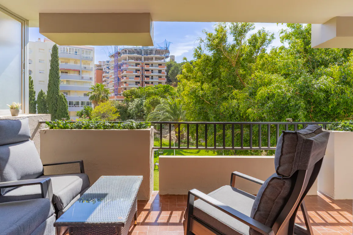 A sunny balcony with gray chairs and a glass table overlooks green trees and city buildings.
