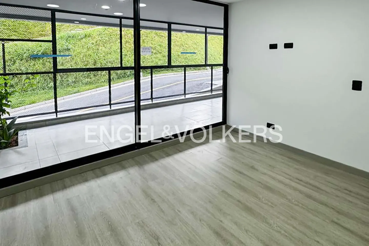 An empty room with light wood floors, white walls, and a large window with a view of a green hillside and road.