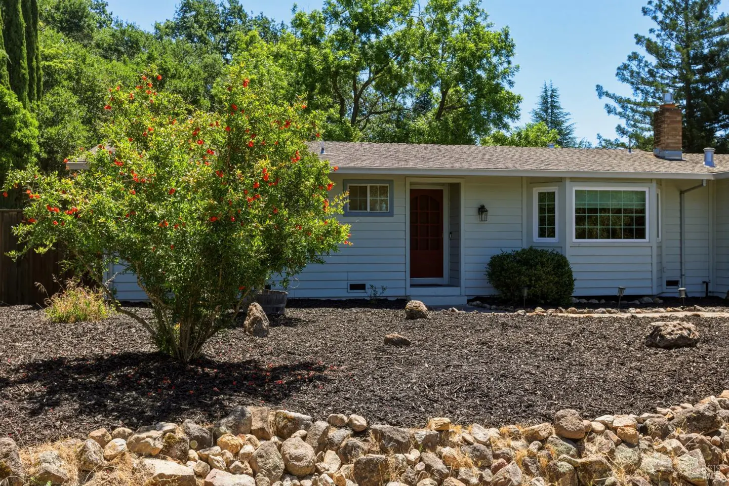 Single-story light gray home with a red front door, surrounded by a rock garden and a flowering tree under a blue sky.