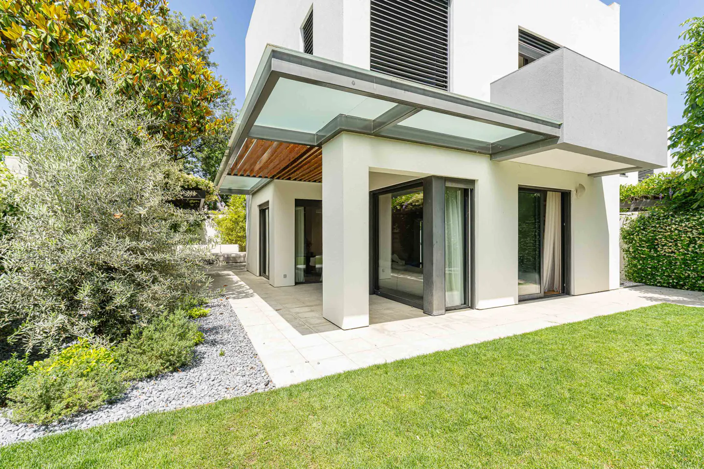Modern white house exterior with a glass-covered patio, green lawn, and lush landscaping on a sunny day.