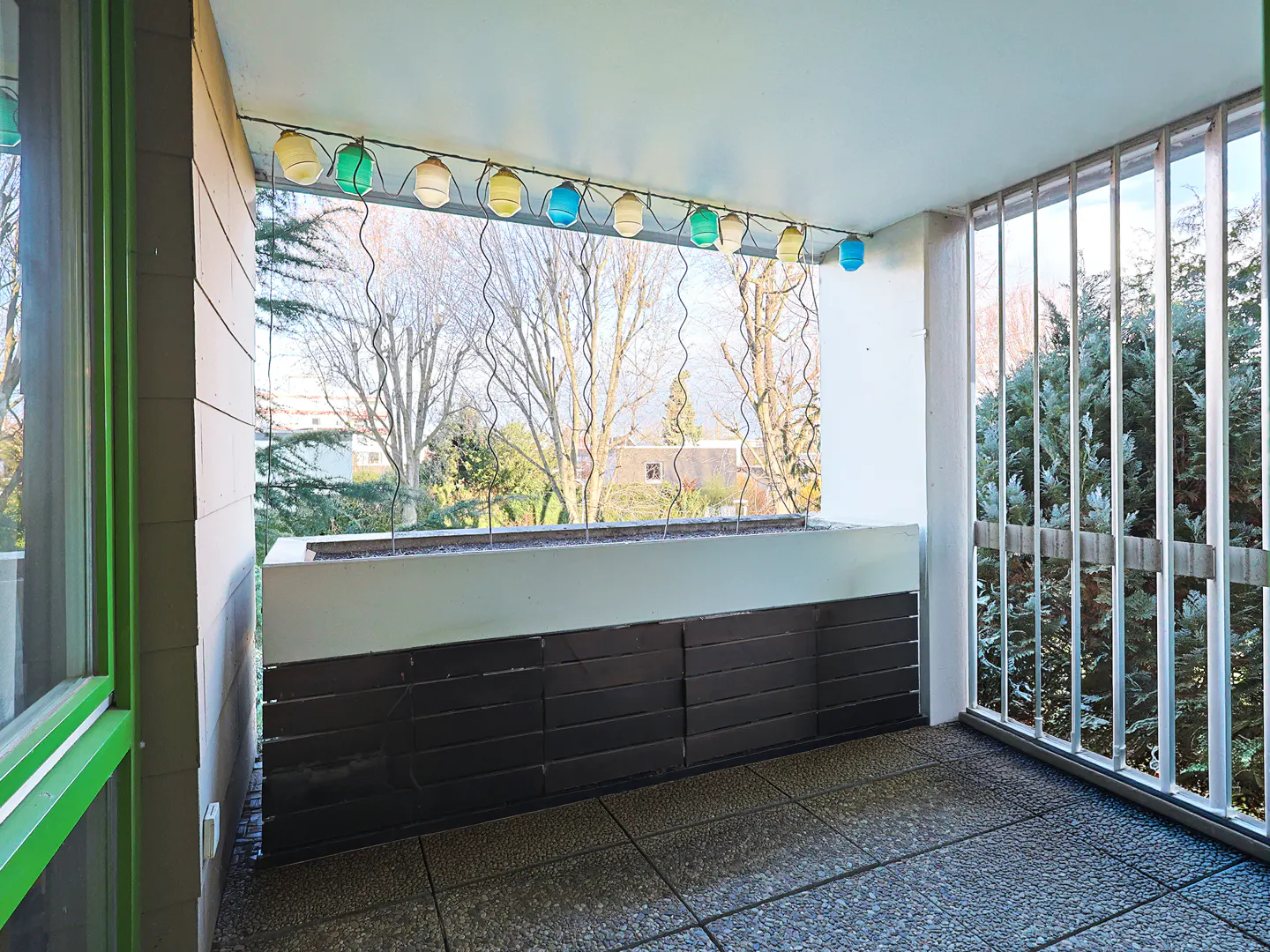 A balcony with a gray tiled floor, a black paneled wall, and a railing with colorful lights. Trees are visible in the background.