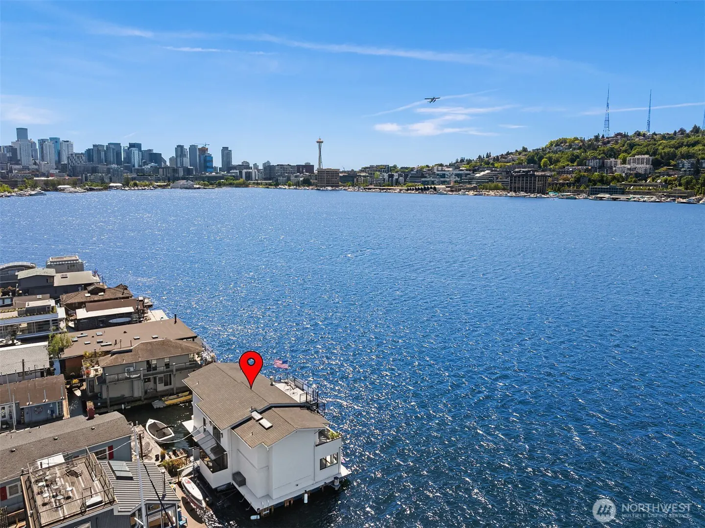 Aerial view of a white houseboat on a blue lake, with Seattle skyline in the background under a clear blue sky. A red pin marks the houseboat.