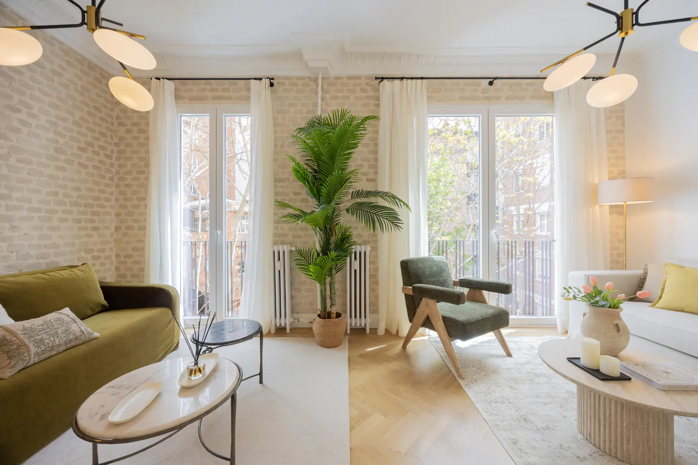 Bright living room with olive sofa, green armchair, and palm tree between two windows with white curtains.