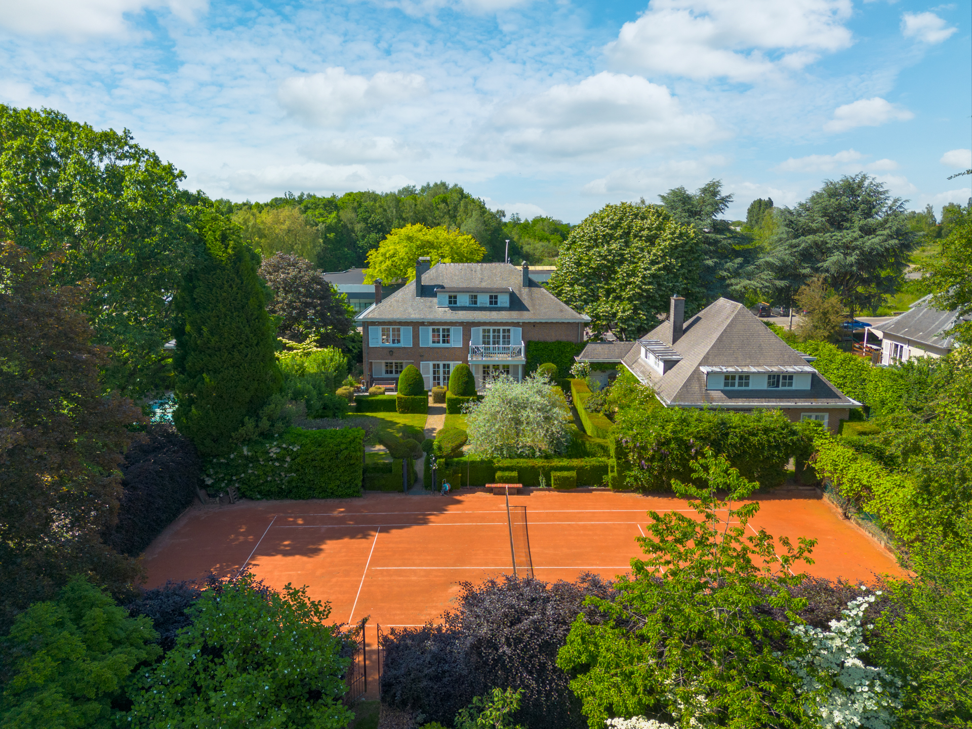 Aerial view of a large house with a clay tennis court in the foreground, surrounded by lush green trees and gardens.