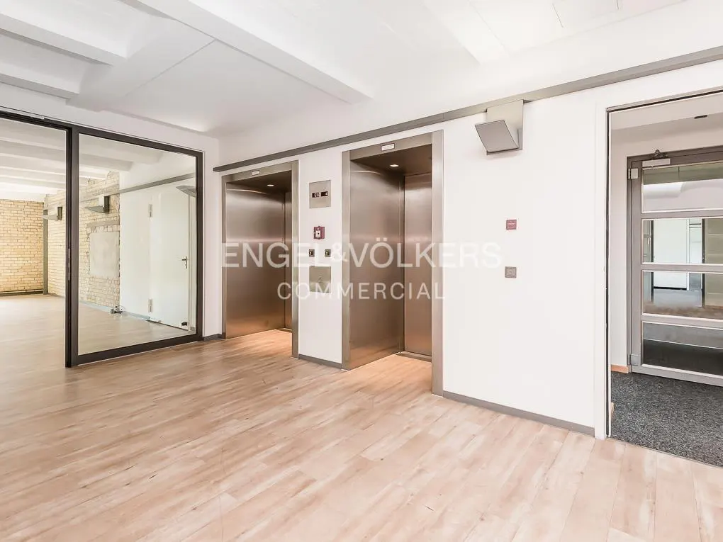 A bright office lobby with two elevators, wood floors, and glass-walled rooms. The Engel & Völkers logo is visible.