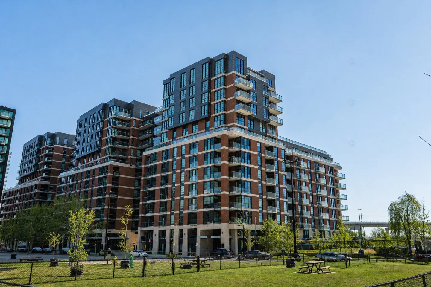 Modern brick apartment building with balconies, viewed from a green lawn with picnic tables under a clear blue sky.