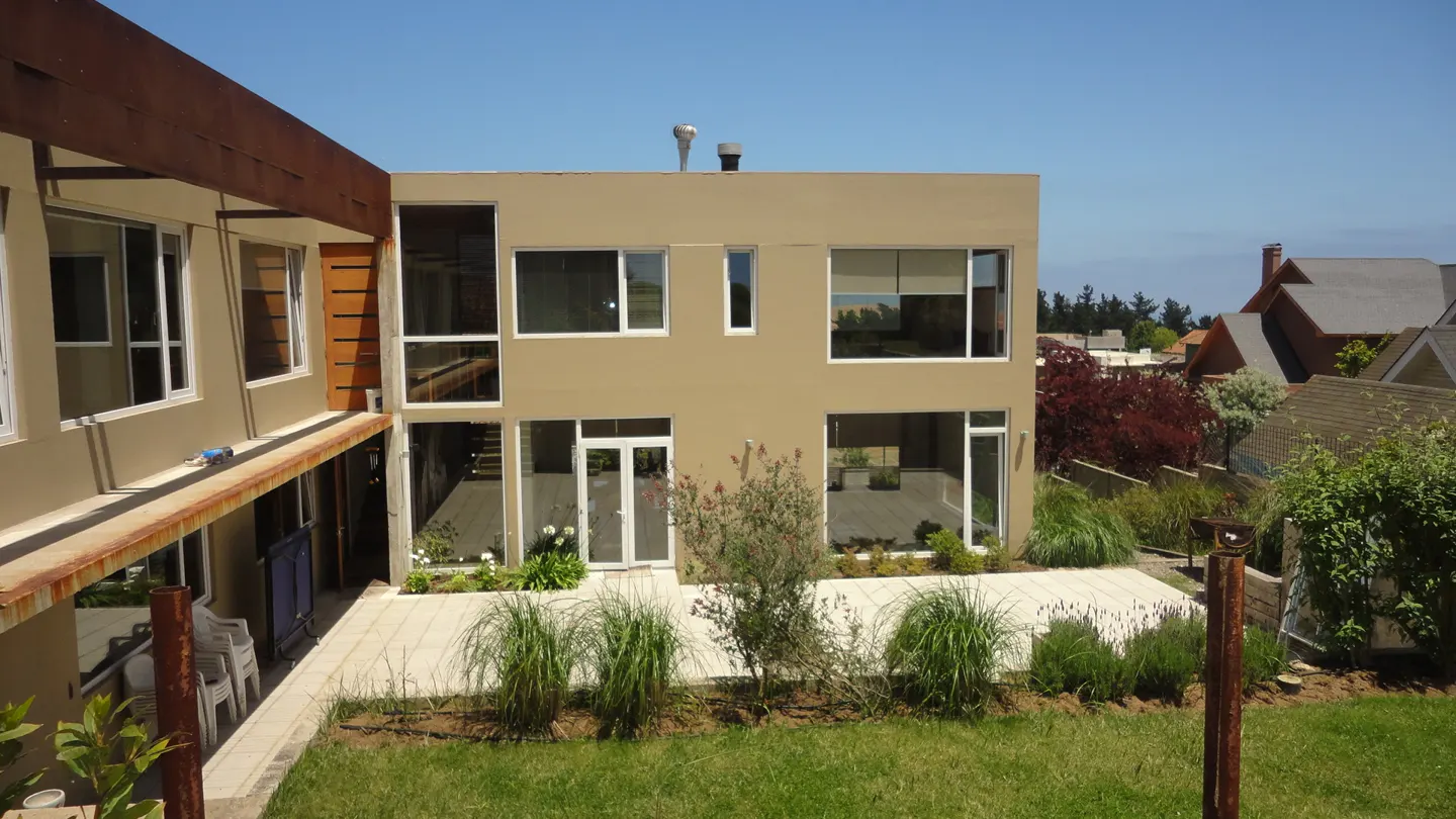 Tan two-story house with white trim and large windows, viewed from the lawn. A patio with plants sits in front of the house.