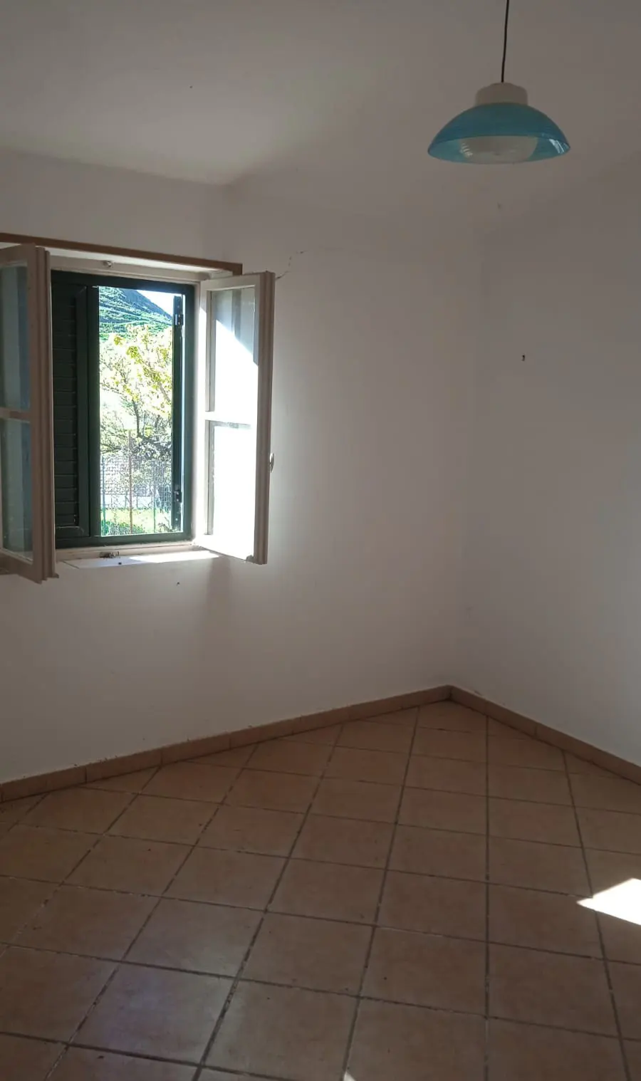 Empty room with white walls, brown tiled floor, and a window with green shutters. A blue lamp hangs from the ceiling.