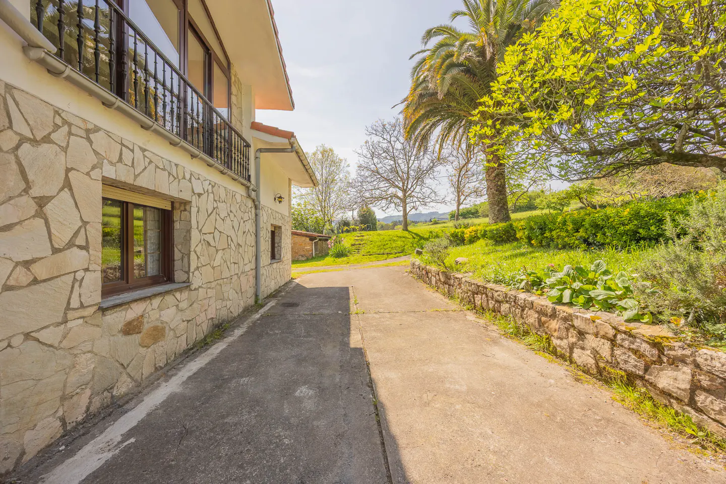 Exterior view of a stone house with a balcony, a concrete driveway, and a green lawn with trees.