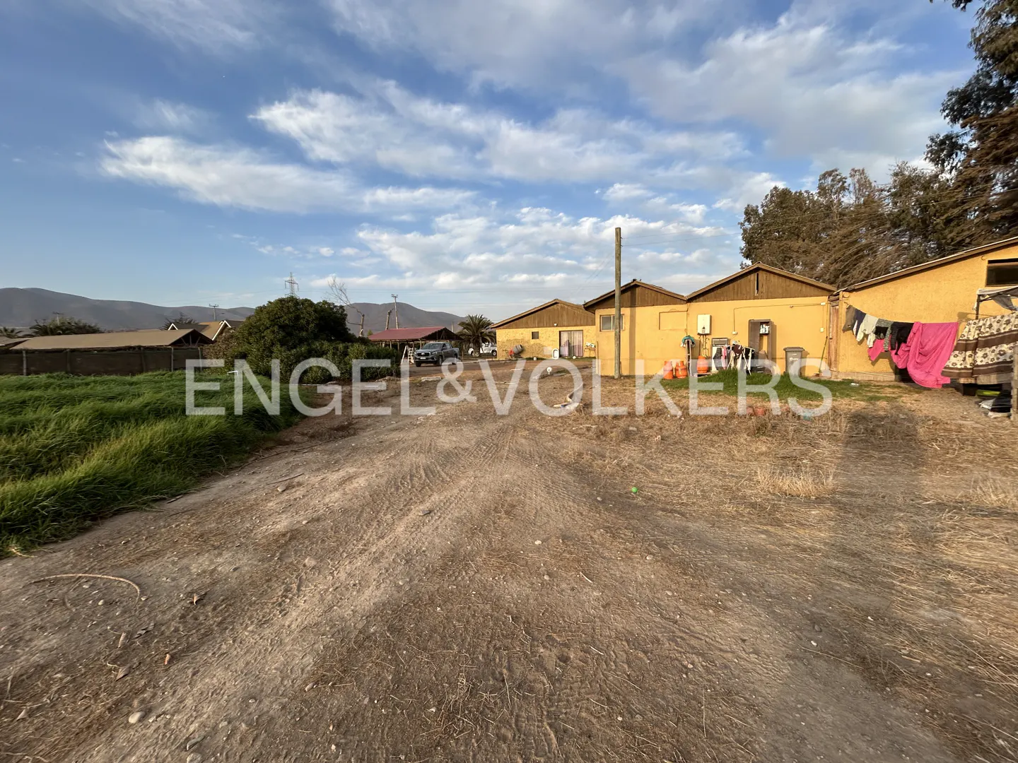 A dirt path leads toward yellow rural buildings under a blue sky with scattered clouds, with laundry hanging outside.