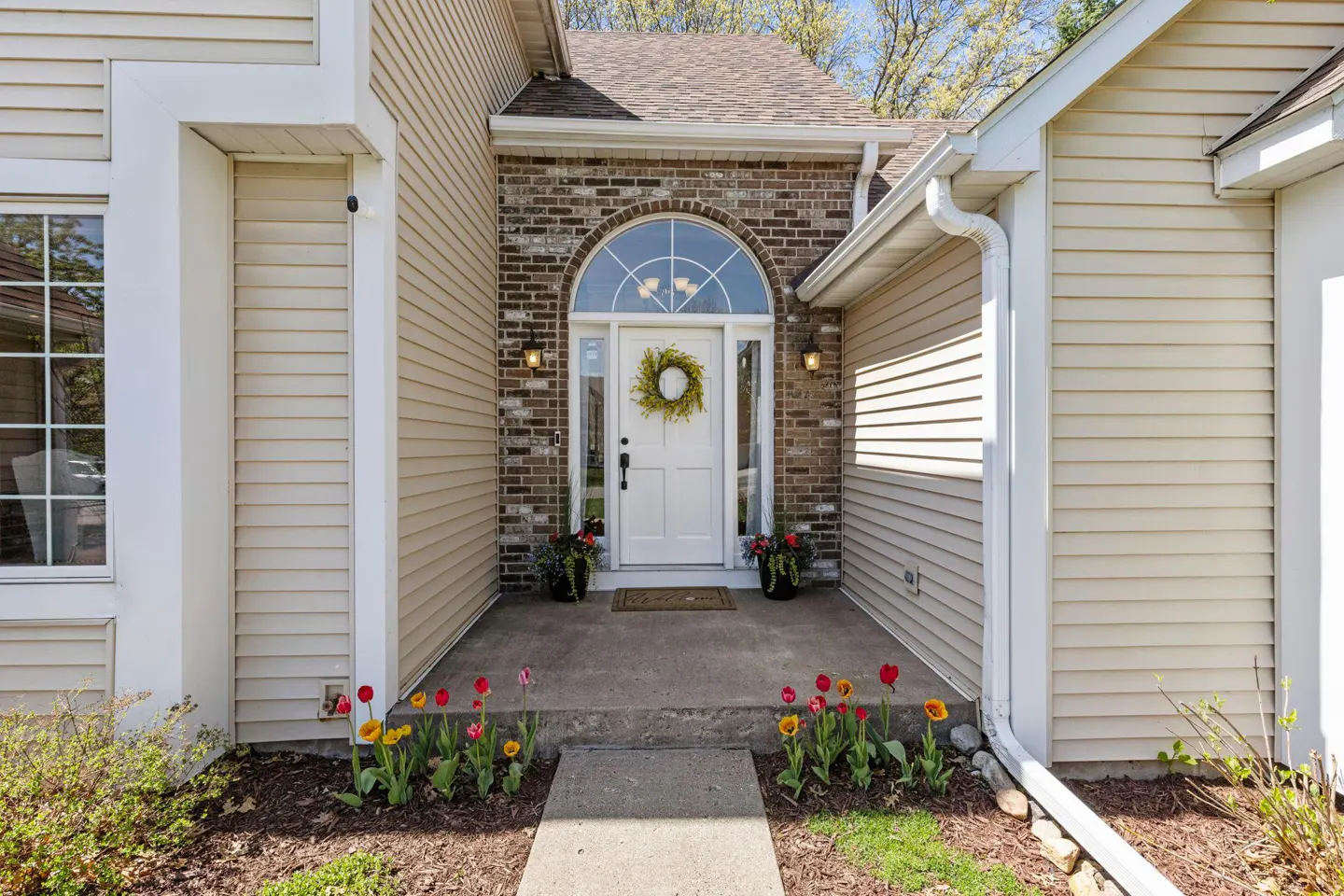 Beige house with a white front door framed by brick, sidelights, and an arched window. A wreath hangs on the door, and tulips line the walkway.