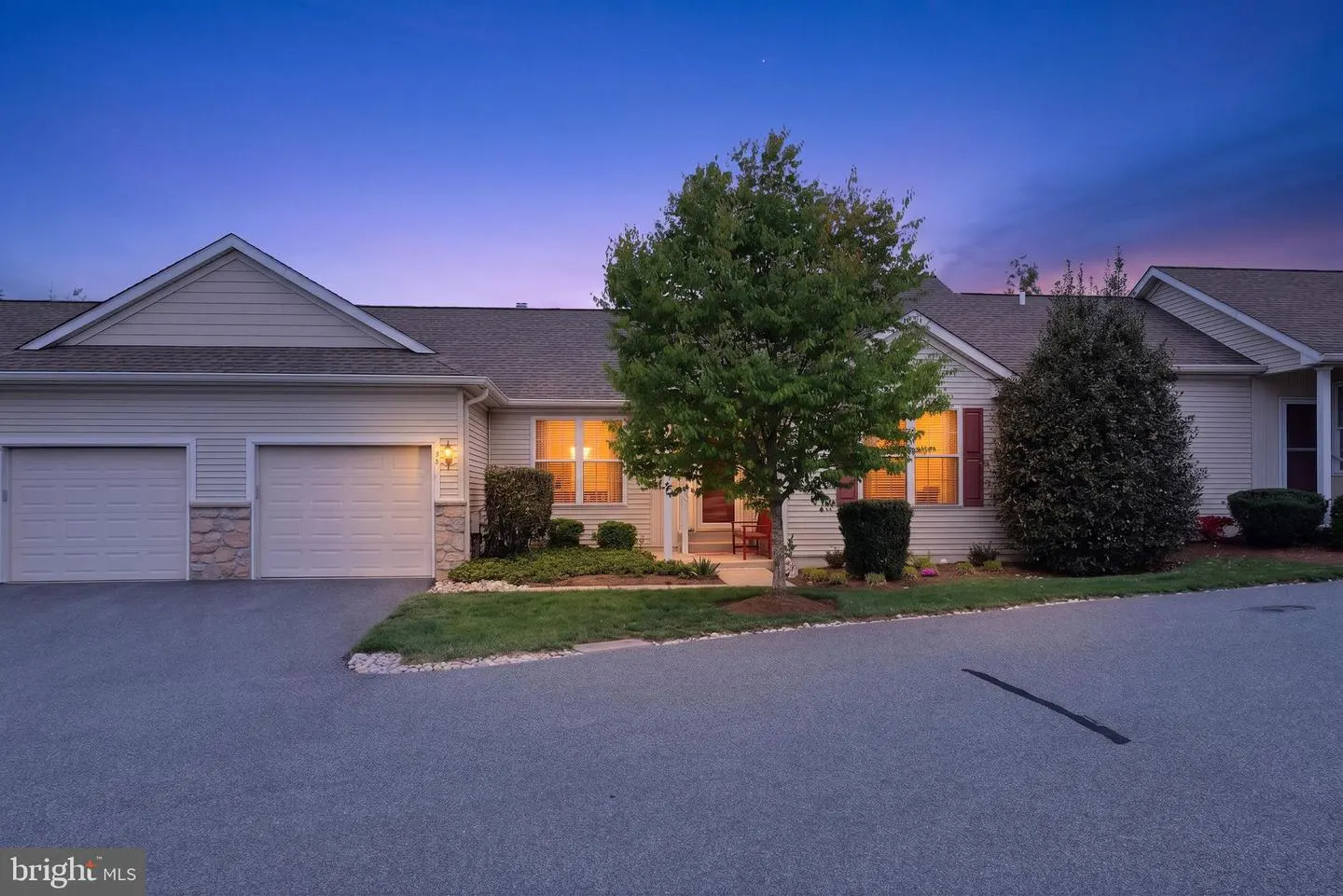 Exterior view of a beige townhouse with a two-car garage and a tree in front at dusk.