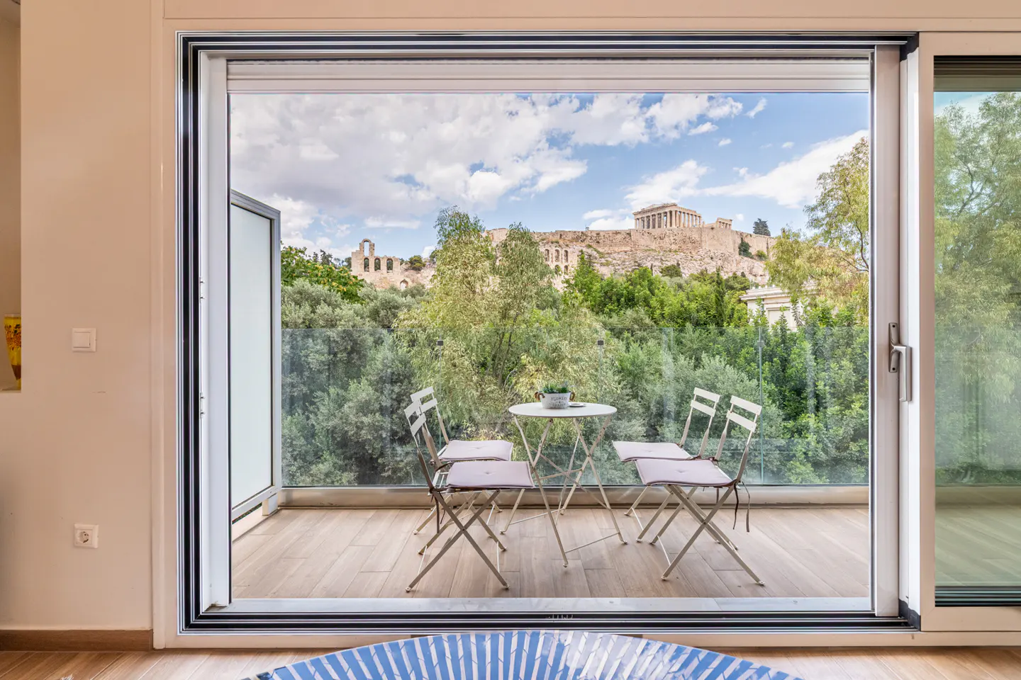 A balcony with a small table and chairs overlooking lush trees and the Parthenon in Athens under a blue sky.