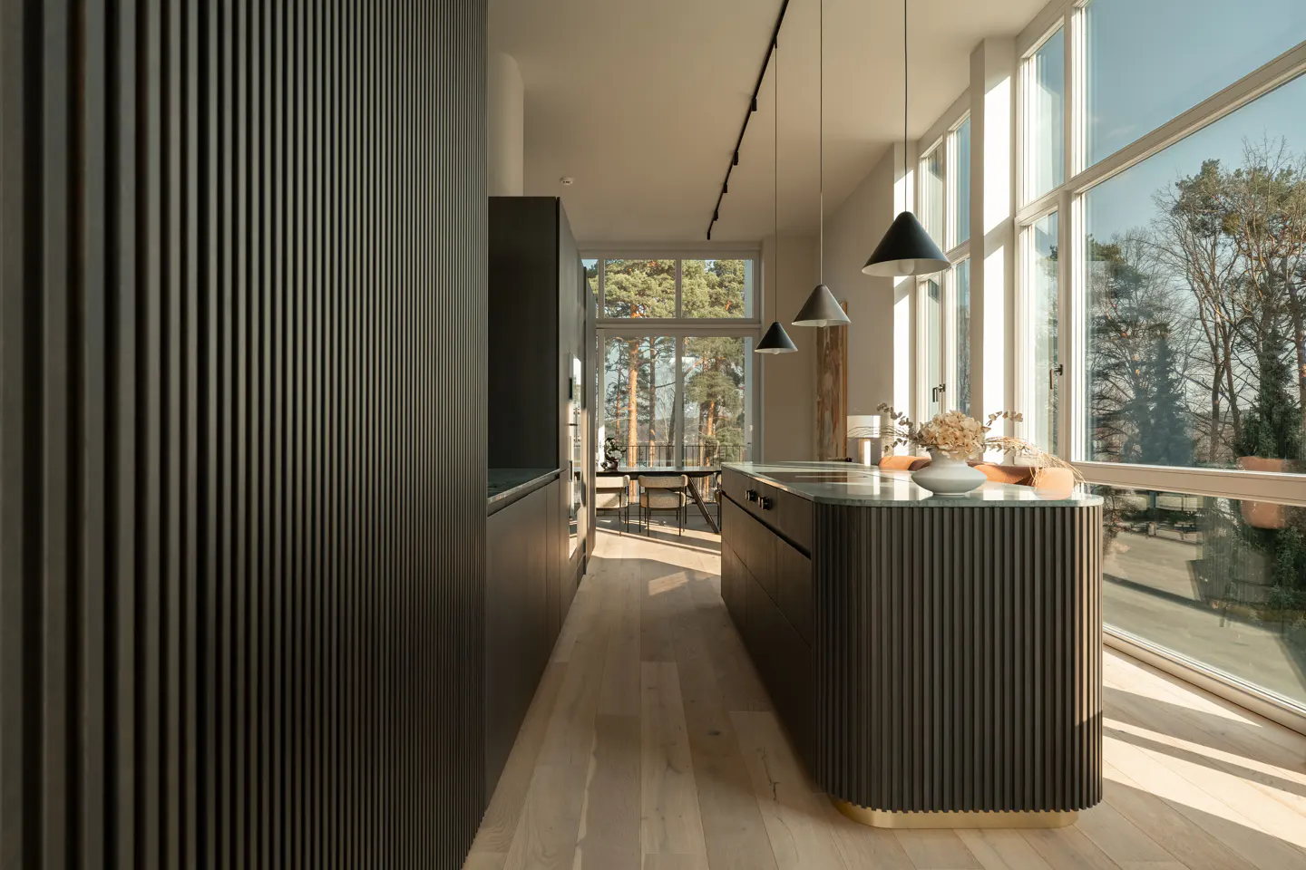 Modern kitchen with dark cabinets, light wood floors, and large windows overlooking trees. Pendant lights hang above the island.