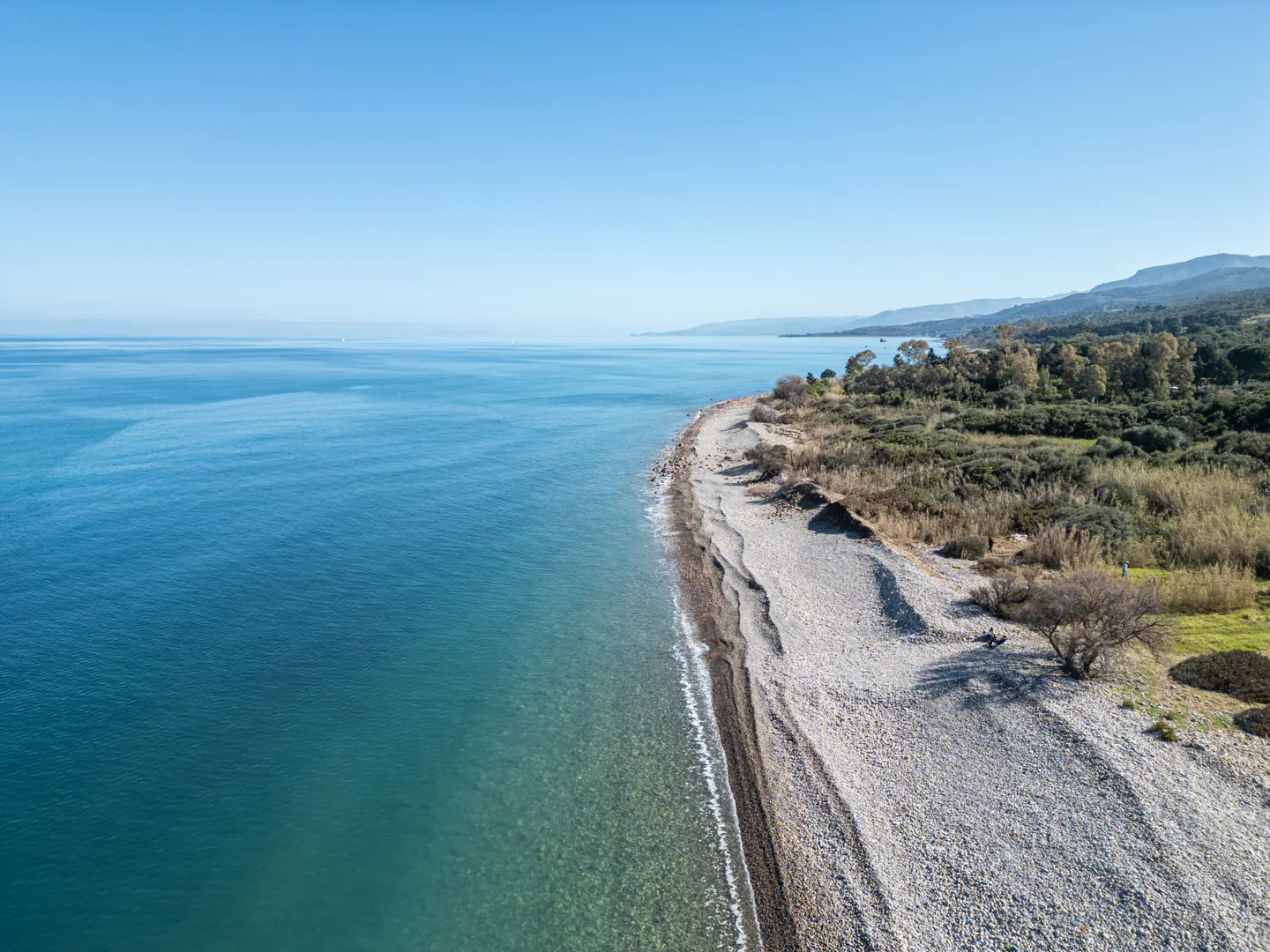 Aerial view of a pebble beach meeting turquoise water under a clear blue sky, with green vegetation and distant mountains.
