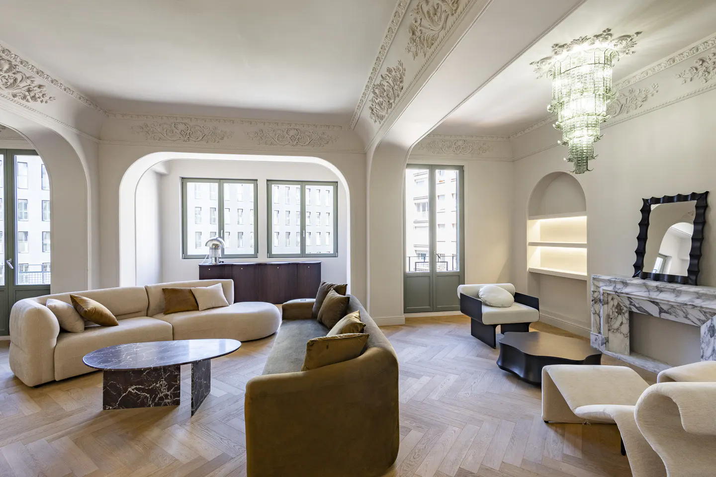 Bright living room with herringbone wood floors, curved sofas, marble fireplace, and ornate ceiling details.