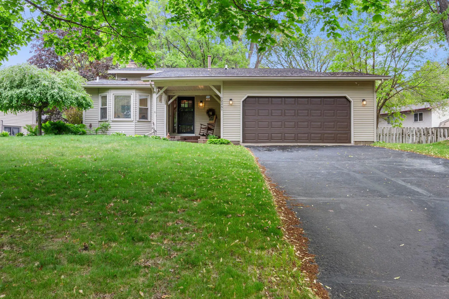 Tan single-story house with a brown garage door and black front door. Green lawn and trees surround the house.