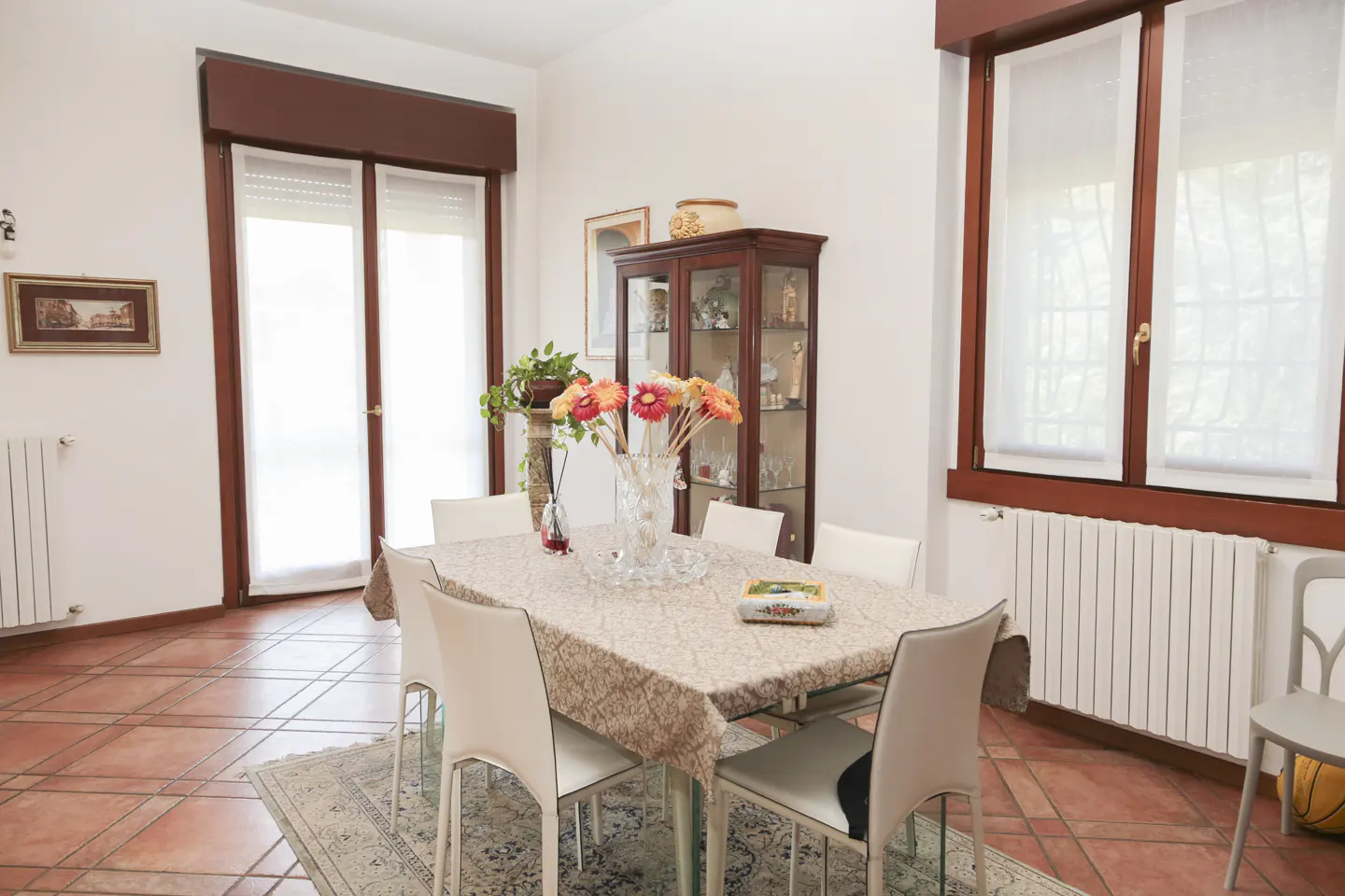 Dining room with a table covered with a patterned tablecloth, white chairs, and a vase of orange flowers. A china cabinet is in the background.