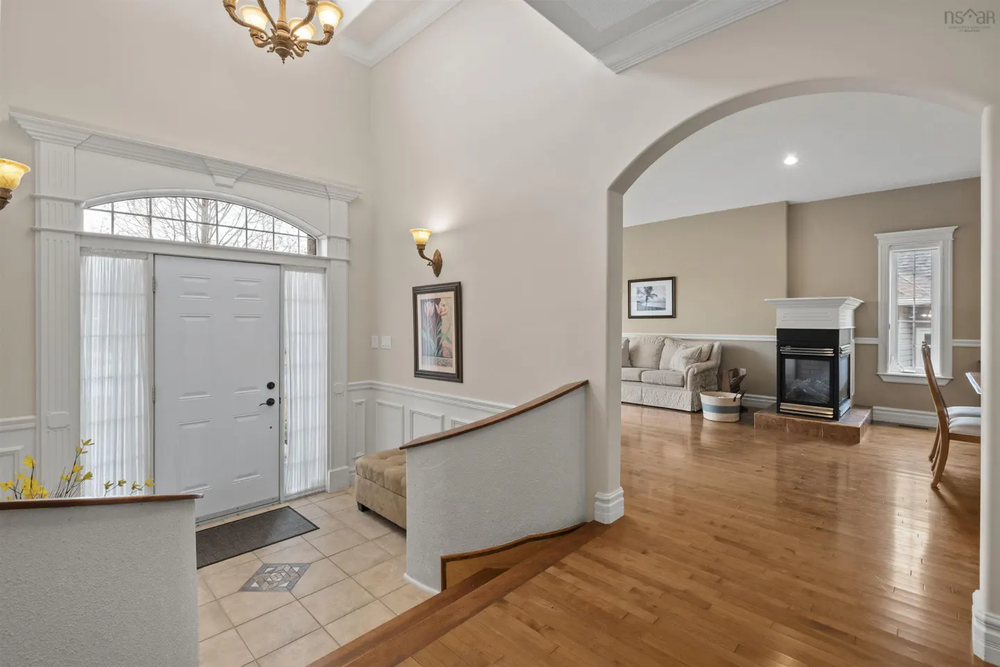 A bright foyer with a white front door, tile floor, and an arched doorway leading to a living room with hardwood floors.