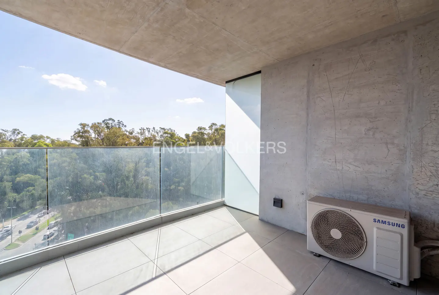 Modern balcony with concrete walls, tiled floor, glass railing, and a Samsung air conditioning unit overlooking trees.