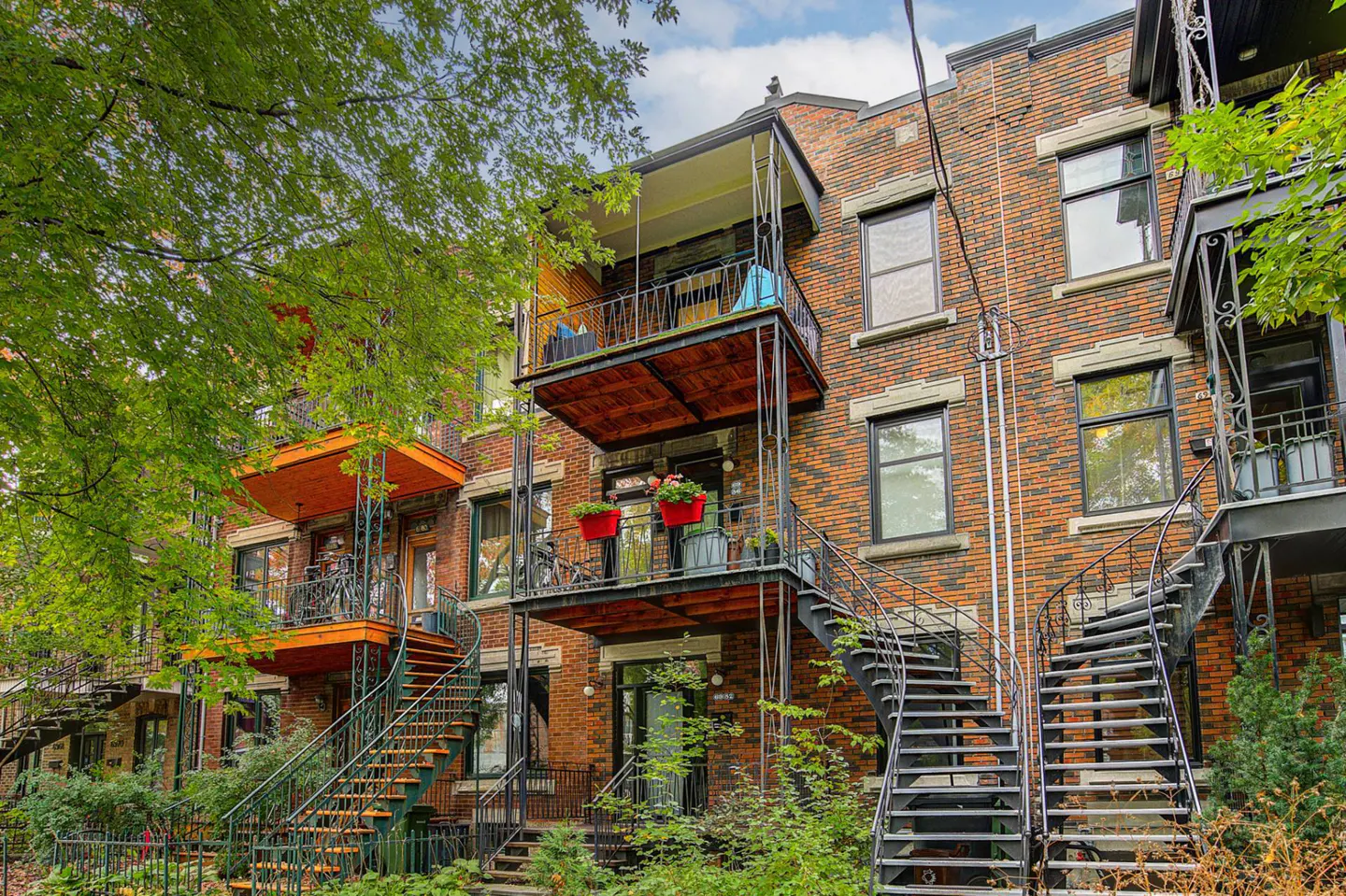 Brick apartment building with black metal stairs and balconies. Green trees frame the building.