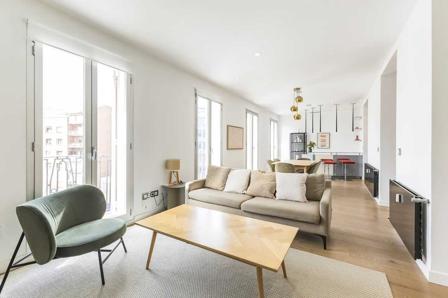 Bright, open-plan living room with a beige sofa, wooden coffee table, and green chair. Large windows let in natural light. Kitchen and dining area visible in the background.
