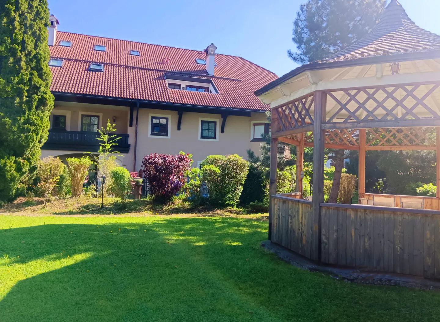 A tan house with a red tile roof sits behind a green lawn and wooden gazebo on a sunny day.