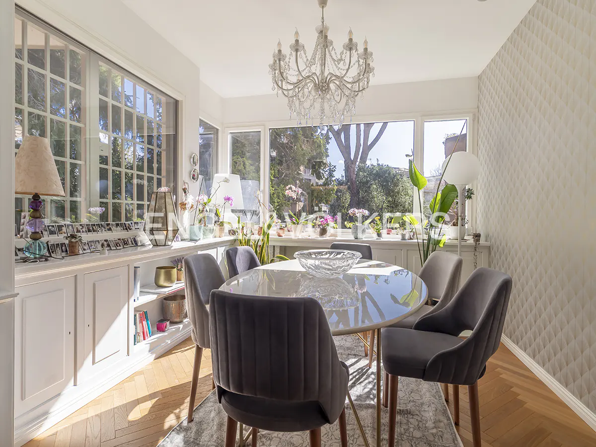 Bright dining room with a round table, six gray chairs, and a crystal chandelier. Large windows overlook a garden.