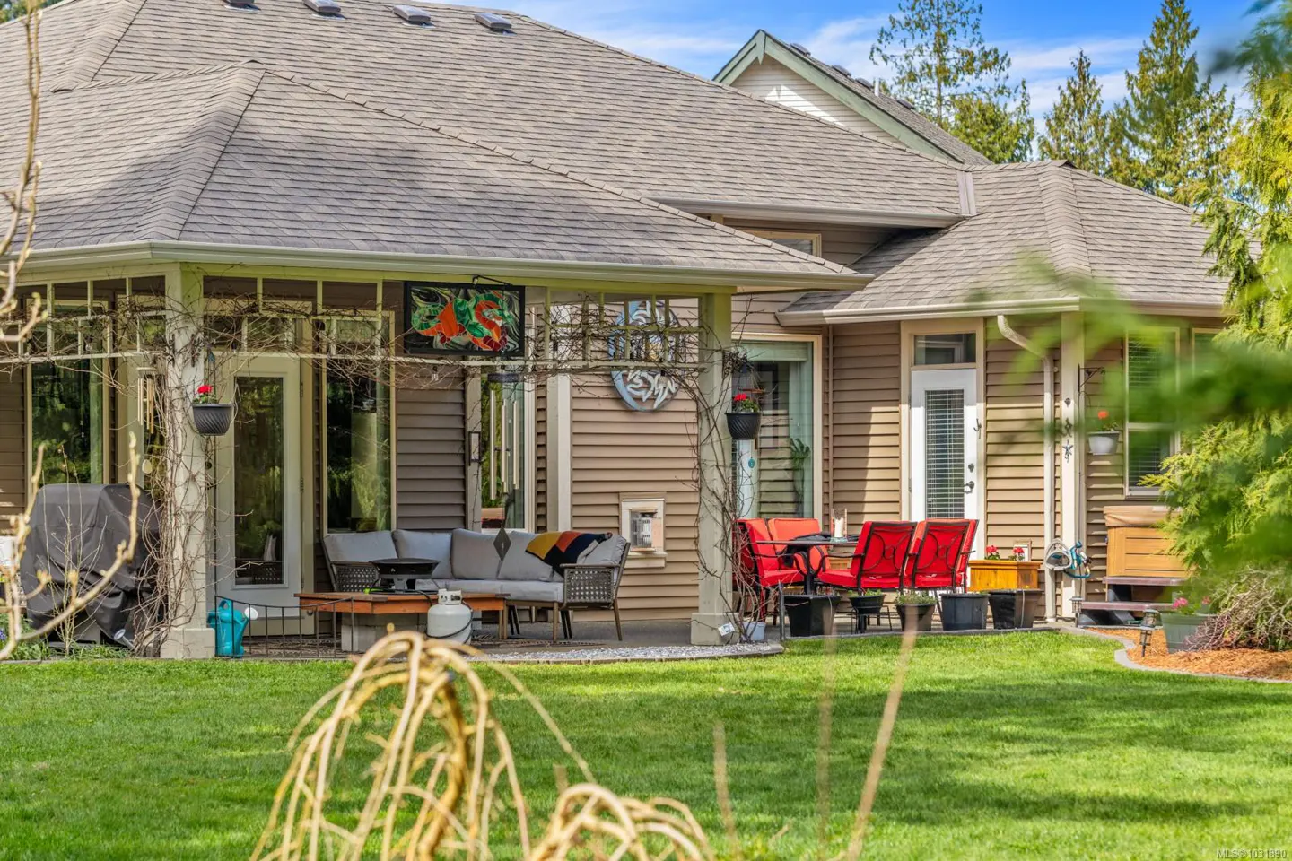 Backyard patio with outdoor seating. A gray sofa and red chairs sit on a concrete patio. Green grass and trees surround the house.
