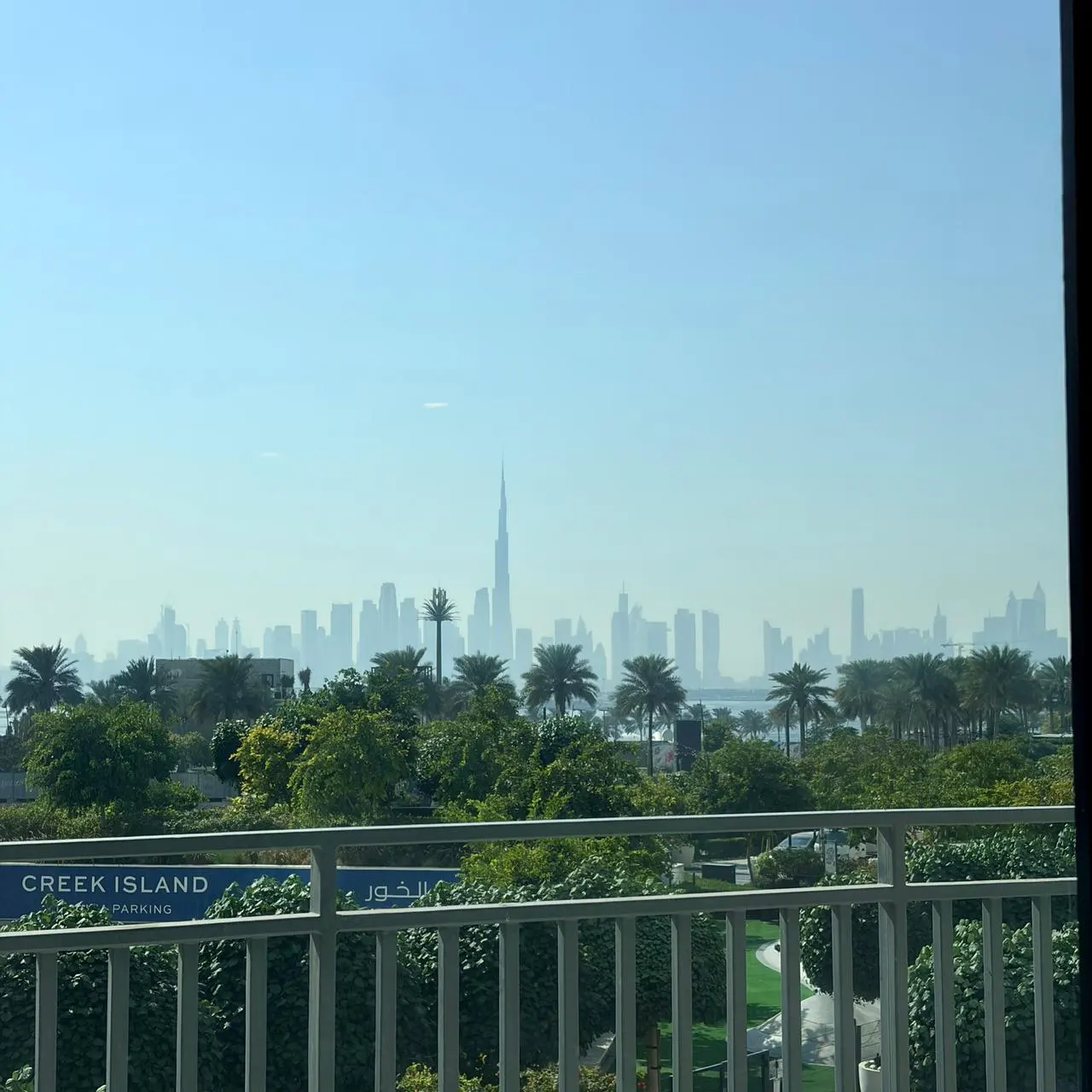 View from a balcony with white railings overlooking green trees and the Dubai skyline in the distance on a hazy day.