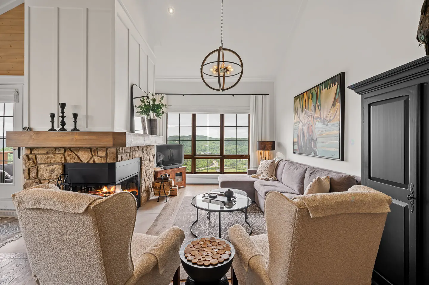 Bright living room with stone fireplace, wood mantle, and two beige armchairs. A gray sofa faces a large window with mountain views.