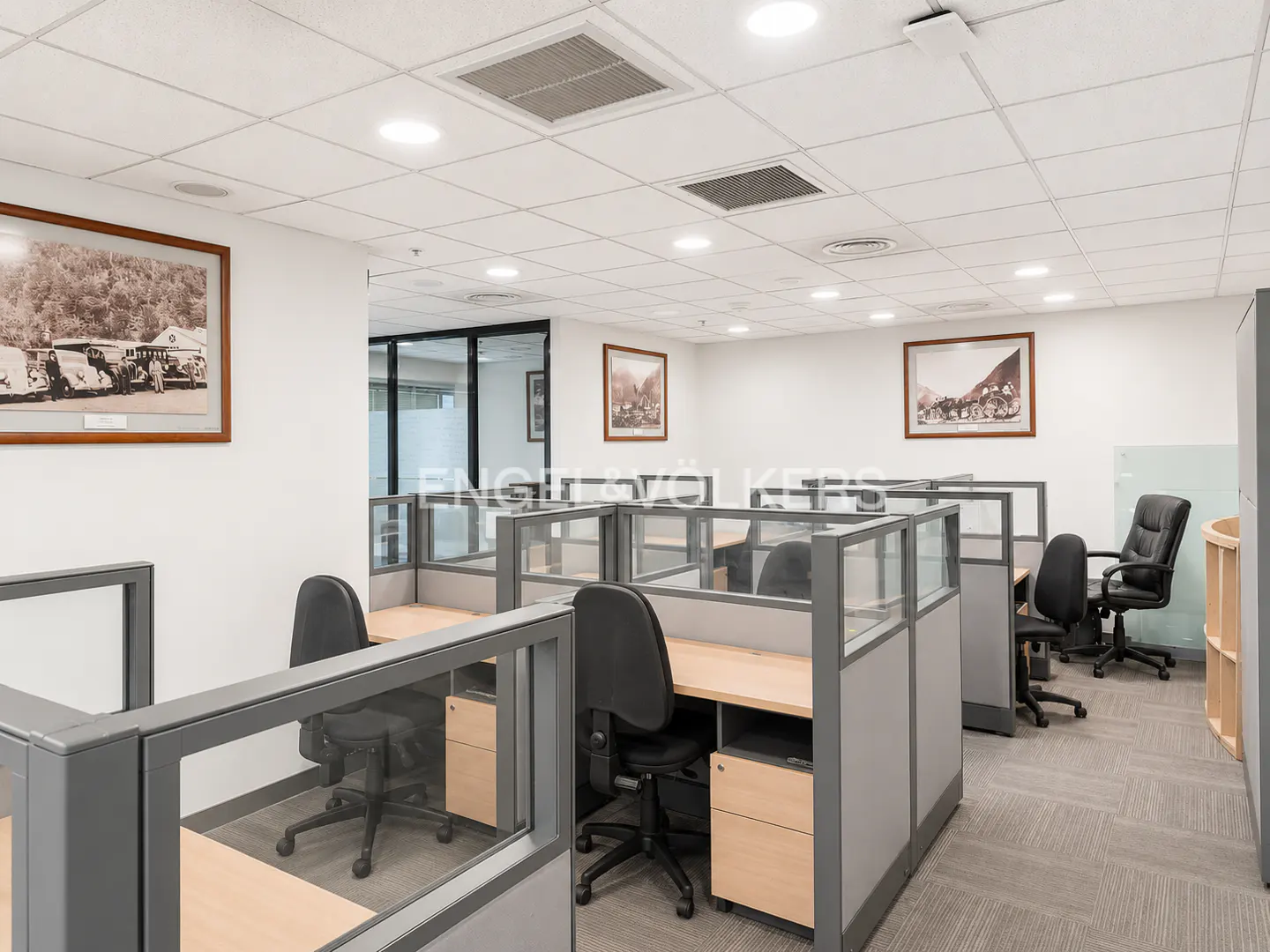 Office space with gray cubicles, black chairs, and light wood desks. Framed pictures hang on the white walls. The floor is gray carpet.