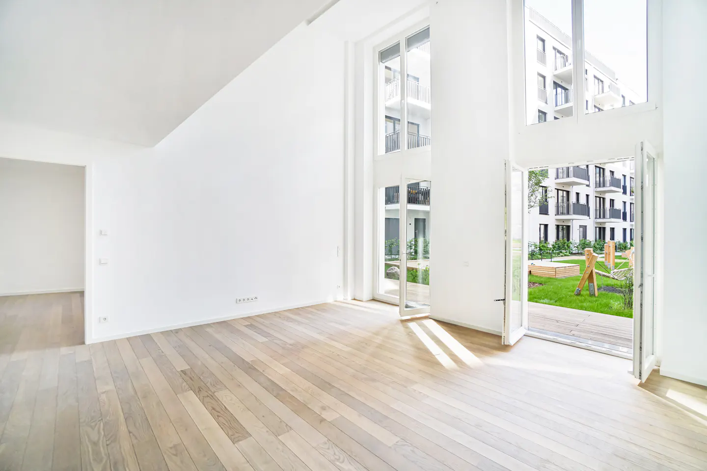 Bright, empty room with light wood floors and white walls. Large windows and doors open to a green courtyard with a modern building in the background.